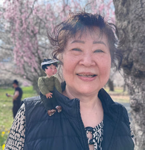 Woman with a tiny grandma puppet on her shoulders in a park with cherry blossom trees